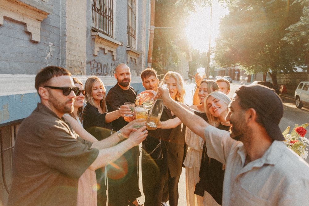 Group celebrates at outdoor wedding reception during golden hour sunset with bright lens flare.