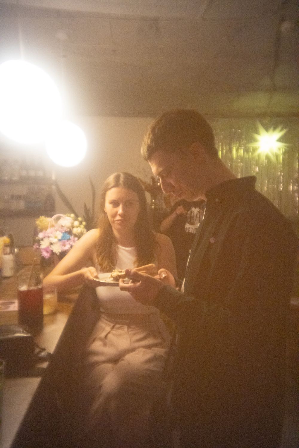 Two people sit at a dimly lit bar or restaurant table looking at something together in warm ambient lighting.