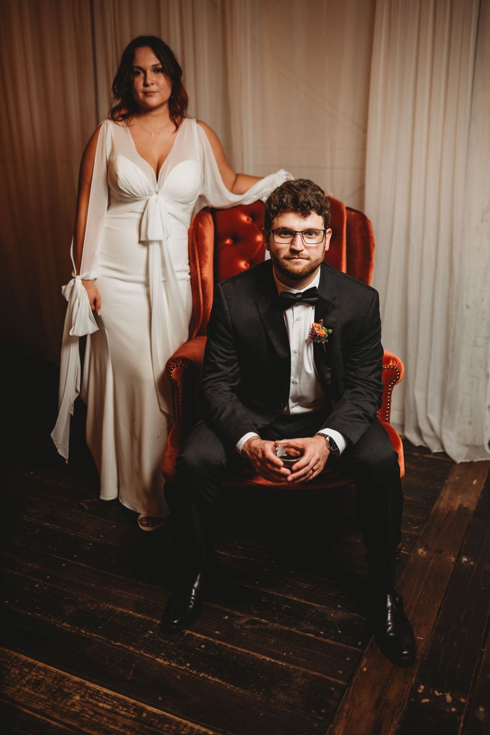 Elegant wedding portrait with vintage red velvet chair against cream backdrop.