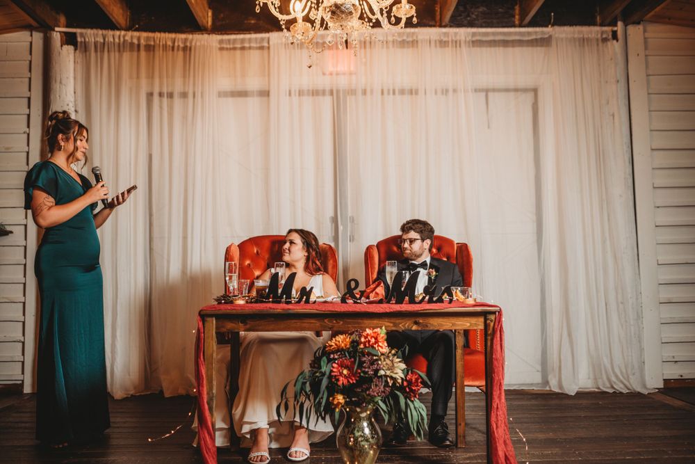 Wedding reception speaker addresses couple at decorated sweetheart table.