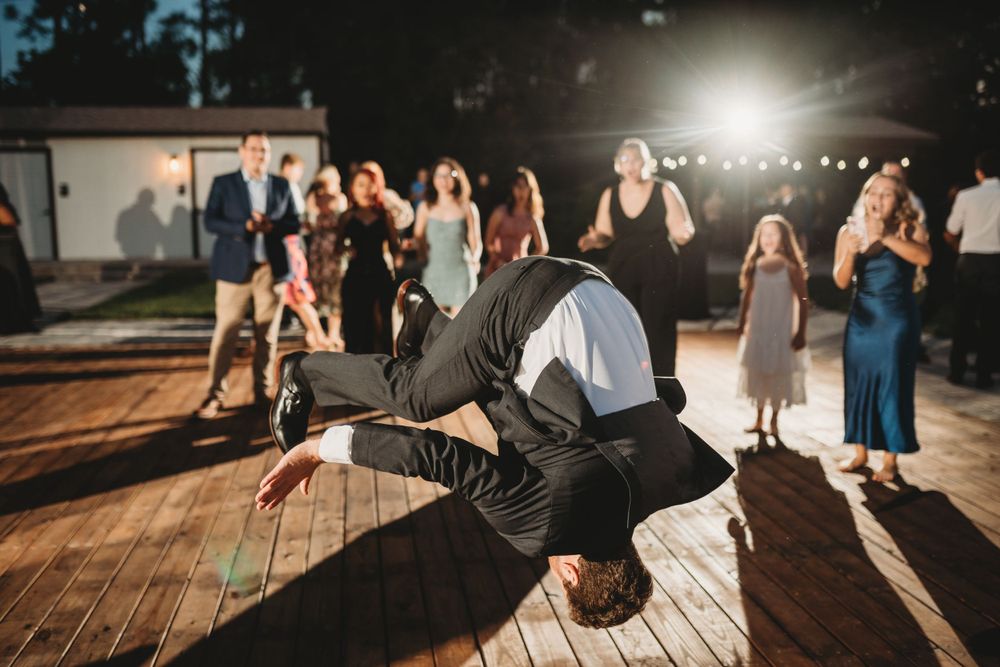 Wedding guest performs impressive breakdancing move on wooden outdoor dance floor at night reception.