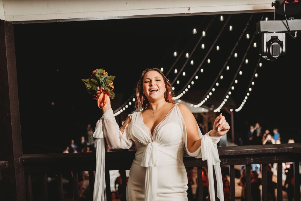 Bride in white dress celebrates with raised arms under twinkling string lights at outdoor reception.