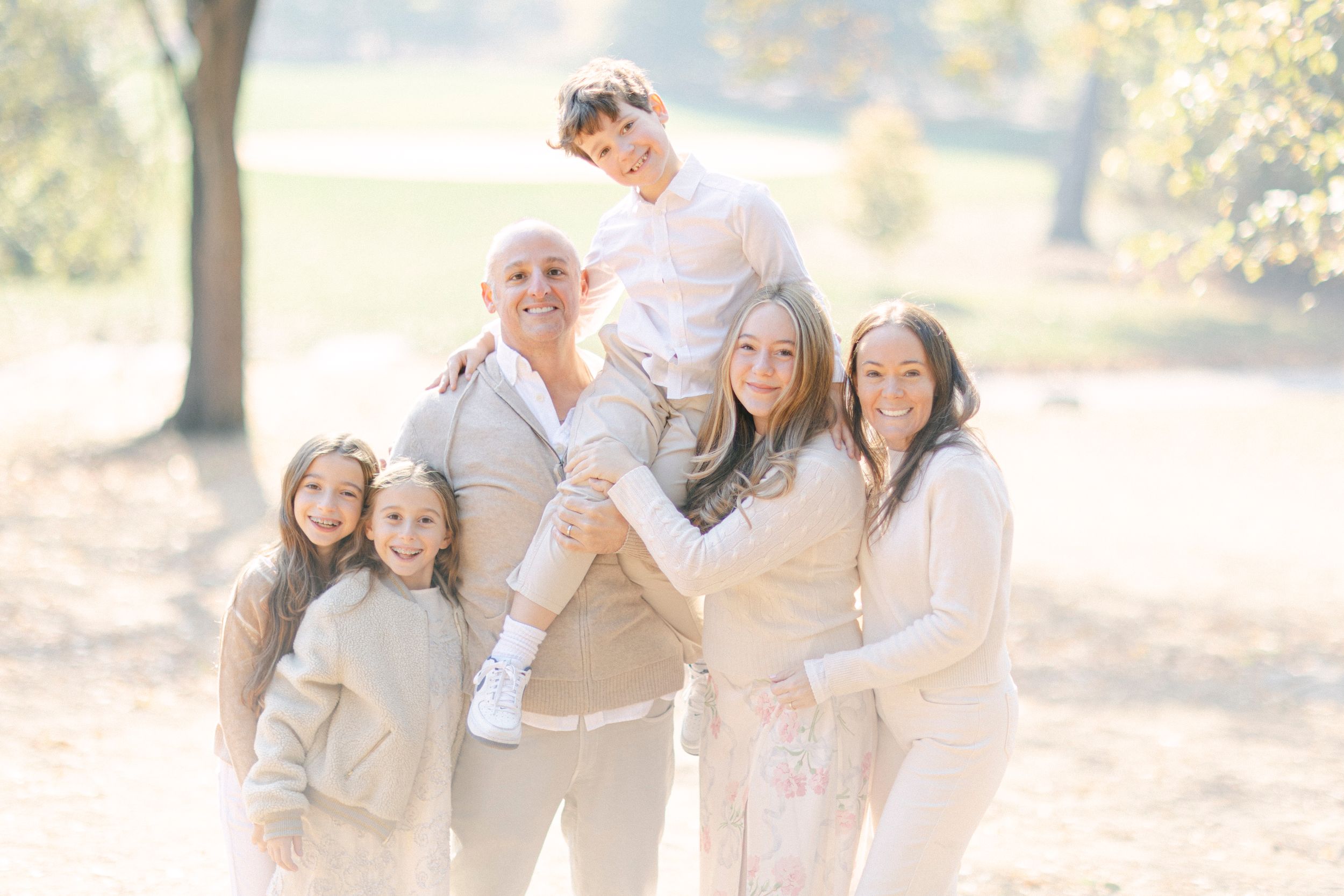 Family portrait outdoors in matching neutral-toned outfits against a sunlit natural background.