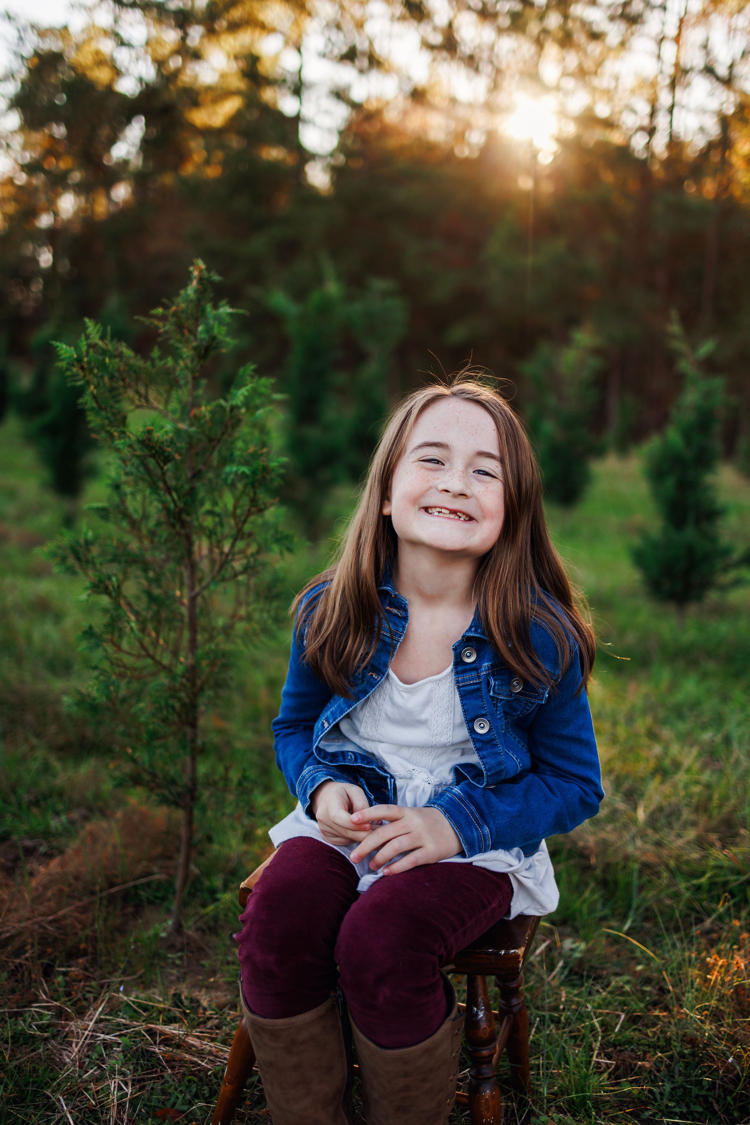 A joyful child in a denim jacket and burgundy pants sits outdoors during golden hour in a natural forest setting.