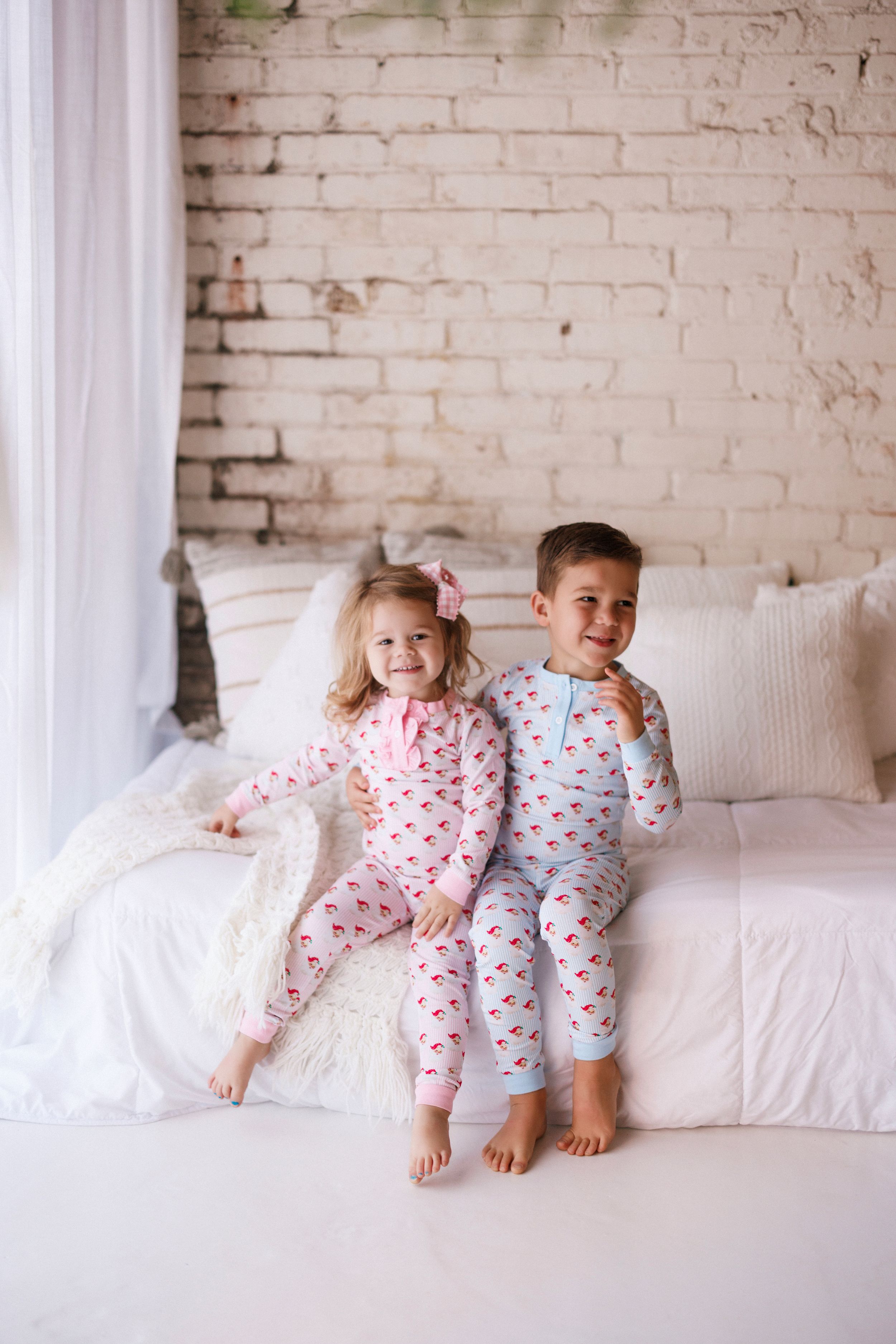 Two children in matching patterned pajamas sit together on a white couch against a brick wall.