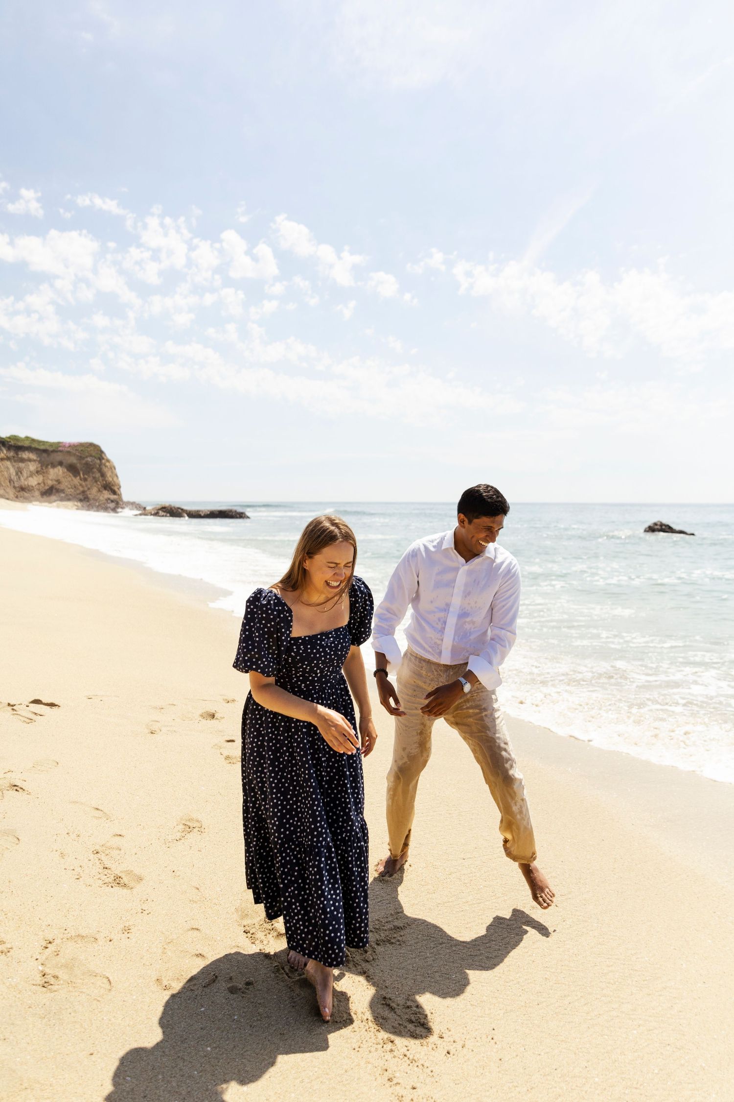 Couple enjoying a romantic moment together on a sandy beach with ocean waves and rocks in the background.