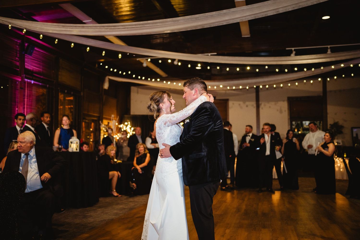 Couples dance together at an elegant wedding reception under twinkling string lights and purple ambient lighting.