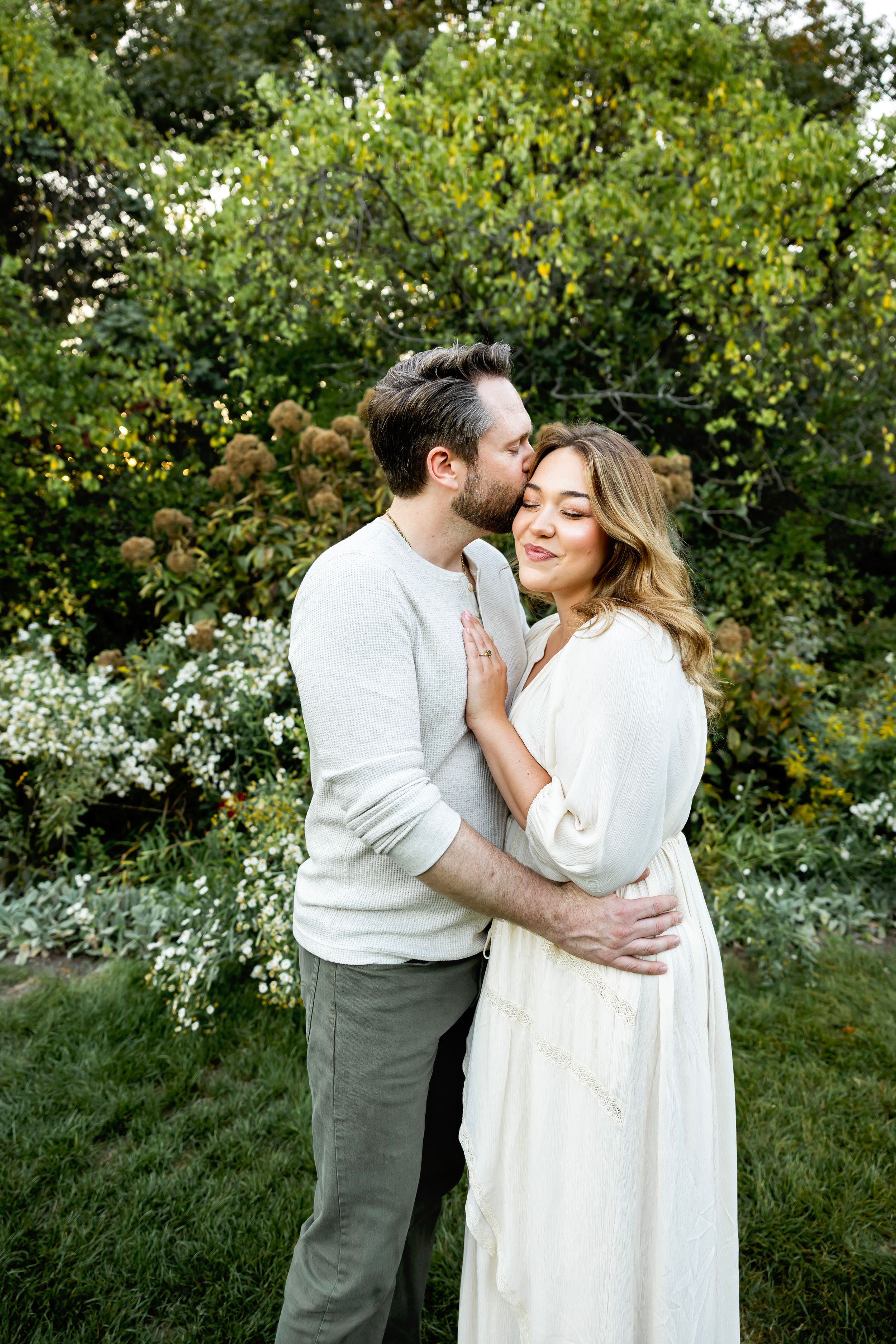 Romantic couple embraces in a lush garden setting wearing white and grey outfits.