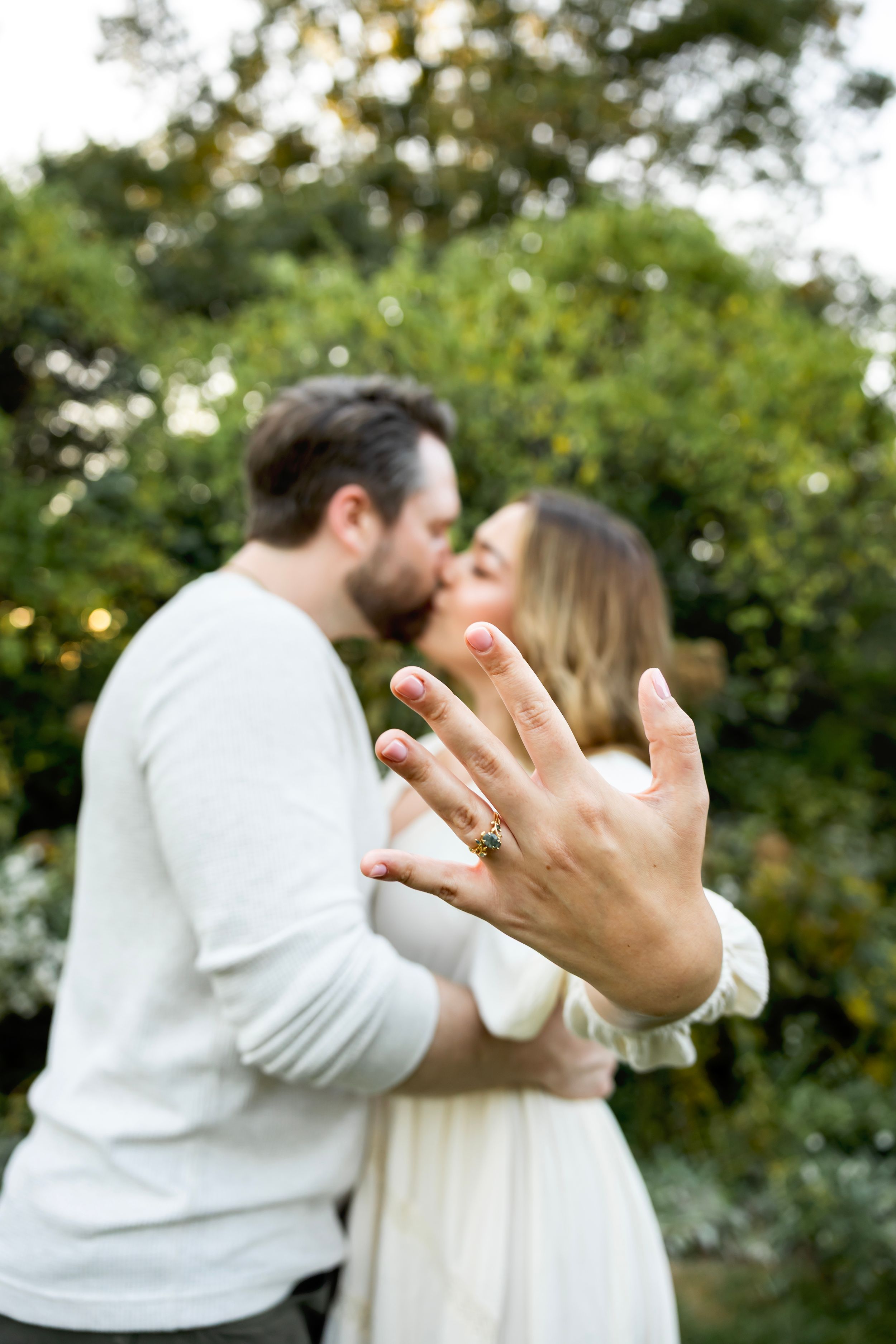 A couple shares a romantic kiss while showing off a ring in an outdoor garden setting.