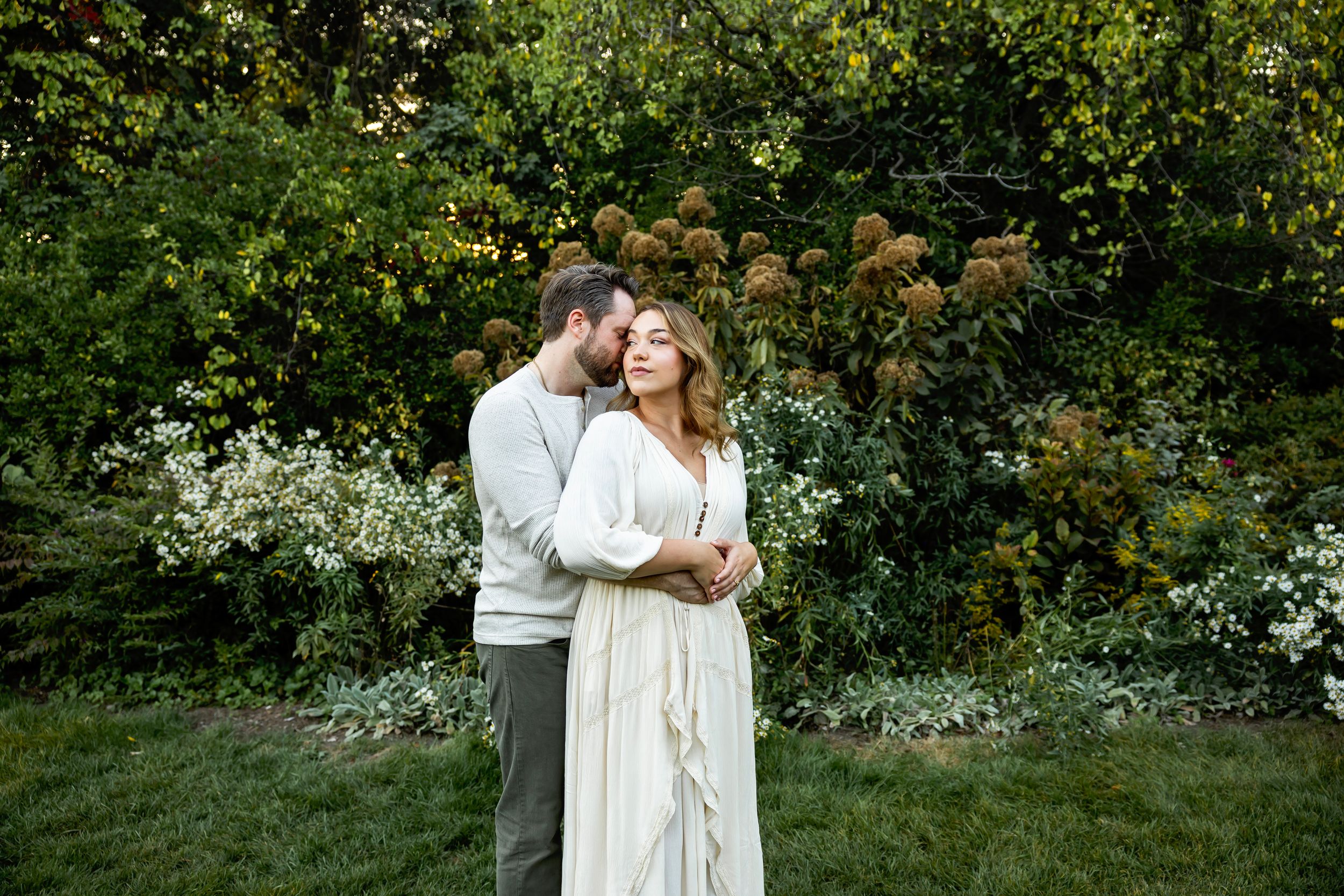 A couple in white outfits embraces romantically in a lush garden setting during a photo session.
