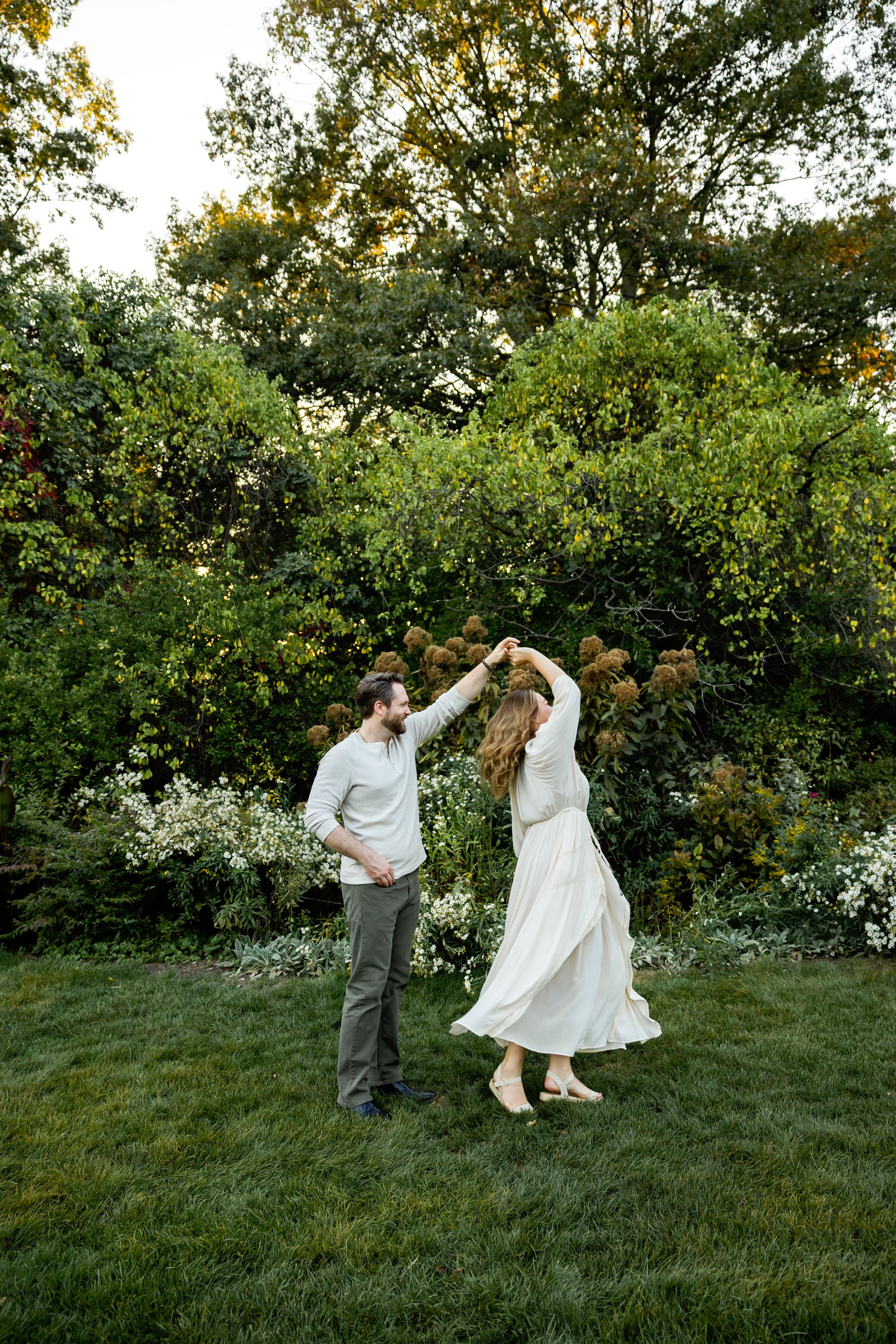 A couple dances playfully on a grassy hill during a golden sunset with trees in the background.