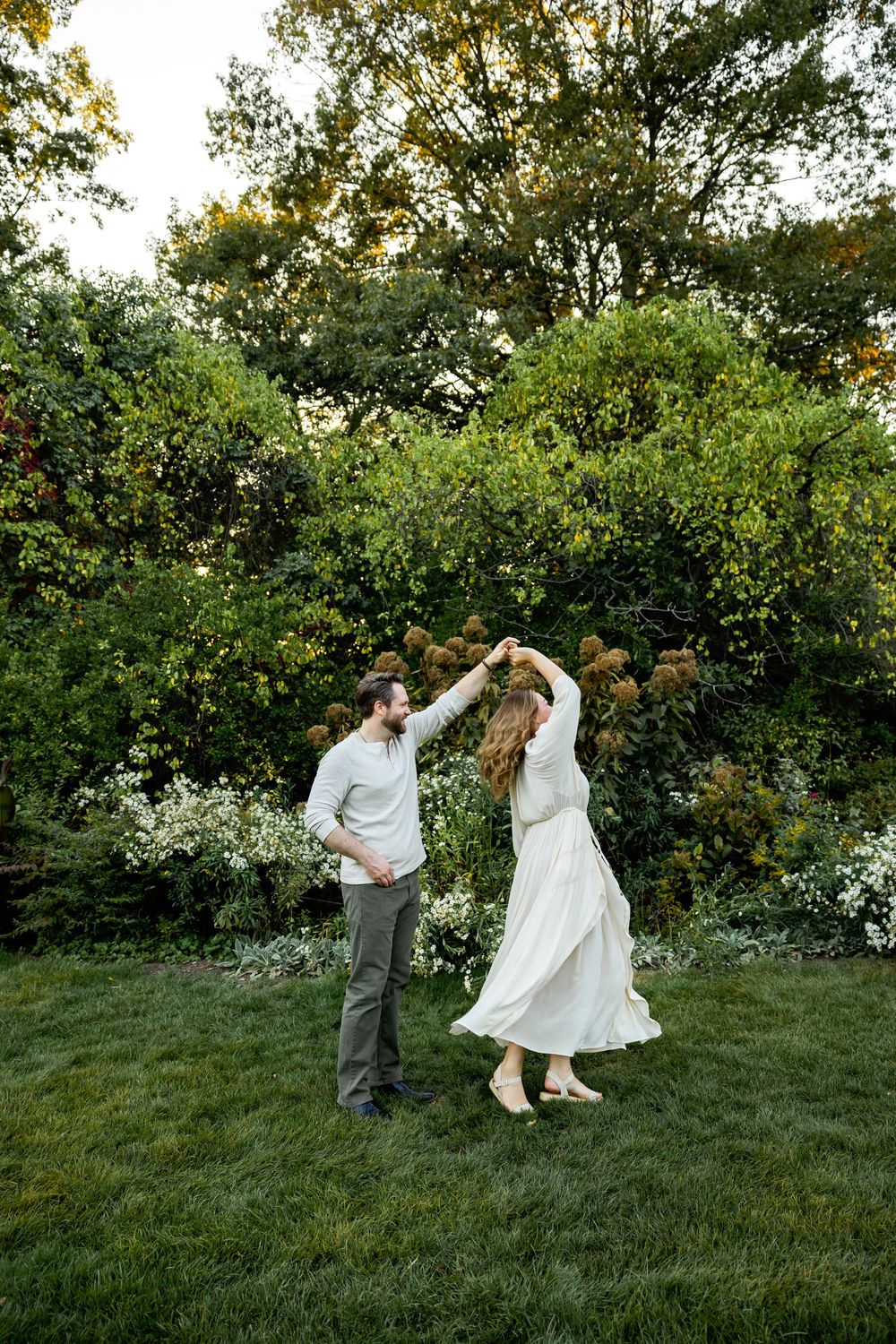 A couple dances playfully on a grassy hill during a golden sunset with trees in the background.
