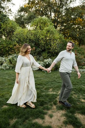A couple in light-colored outfits dances playfully together on grassy lawn during golden hour.