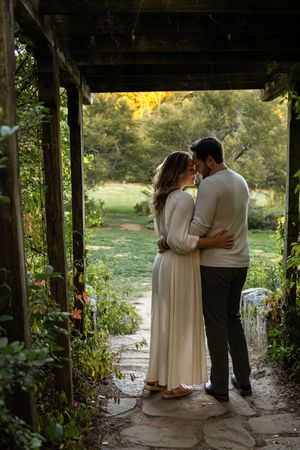 A couple shares an intimate moment under a rustic wooden pergola surrounded by lush greenery and golden sunlight.