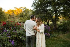A romantic couple embraces in a garden at sunset with colorful autumn foliage and purple flowers in the background.