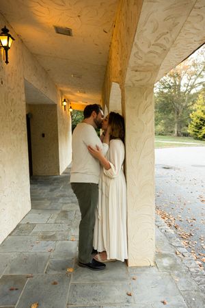 Romantic couple embraces in a white stone colonnade walkway with autumn leaves scattered on the ground.