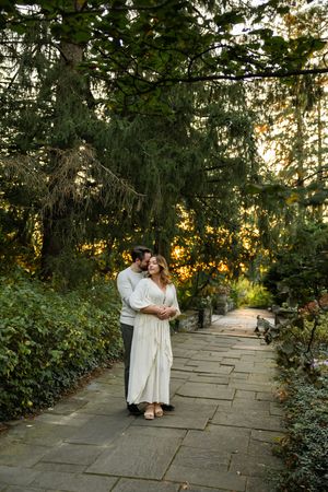 Romantic couple embraces in a lush garden setting with stone pathway and golden sunset light filtering through trees.
