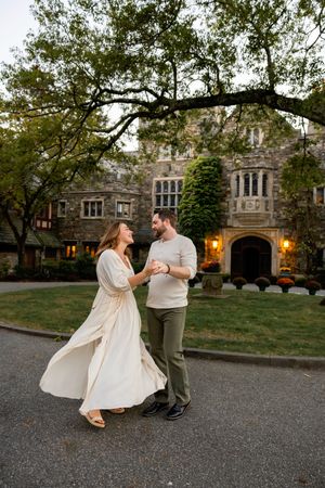 Dancing couple twirls playfully on gravel path in front of gothic stone building at sunset.