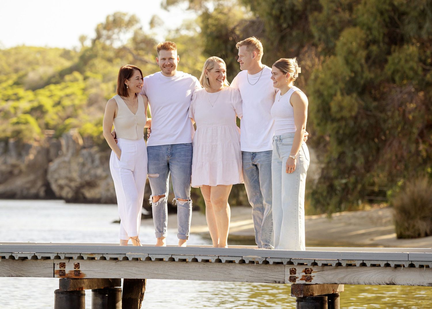 Family wearing white and grey outfits walks together on wooden pier by scenic coastal cliffs at sunset.