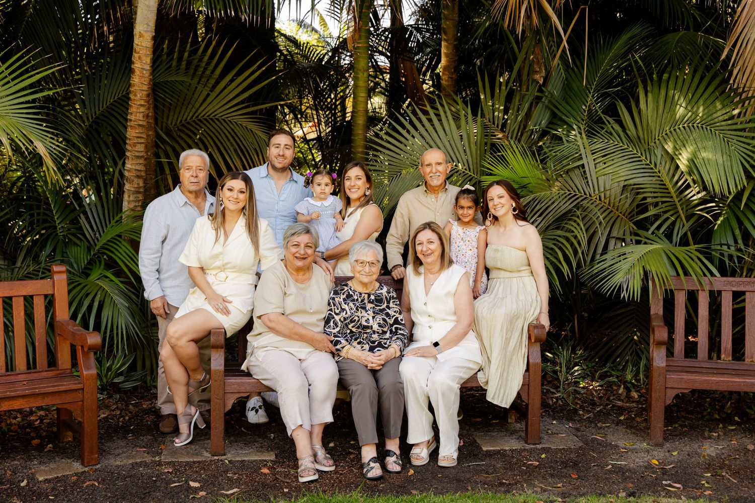 A group in coordinated white and beige outfits poses together on wooden benches surrounded by tropical palm trees.