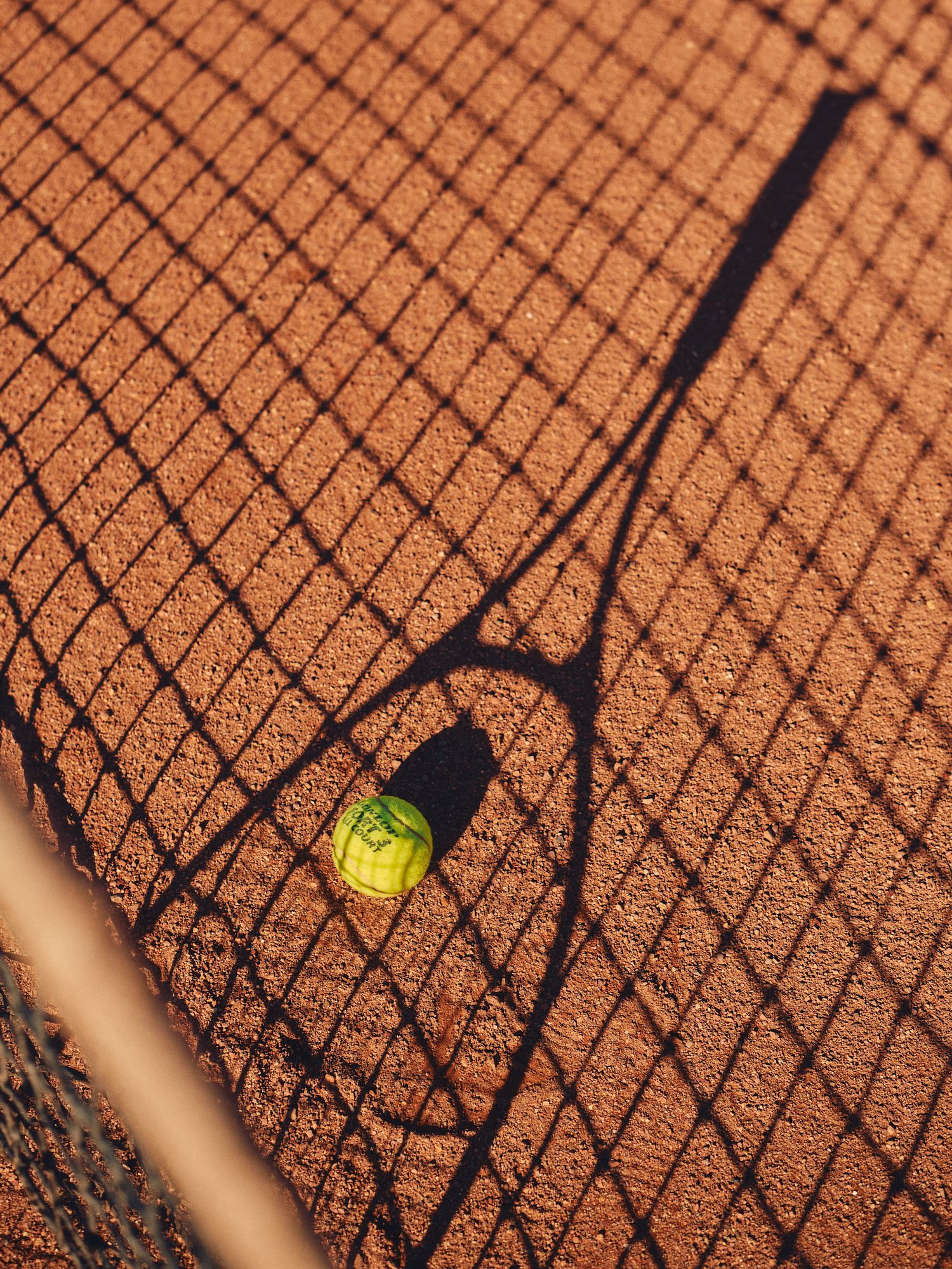 Tennis ball casting dramatic shadows on a clay court surface through a chain link fence.