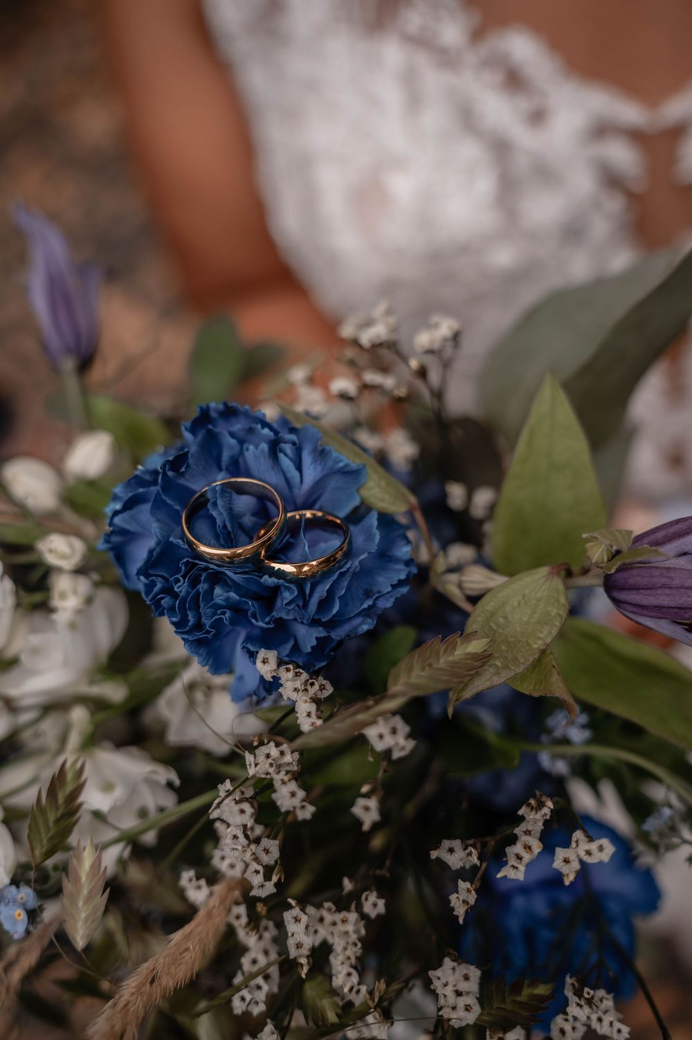 Wedding rings rest on vibrant blue flowers with delicate white baby's breath and greenery in a bridal bouquet.
