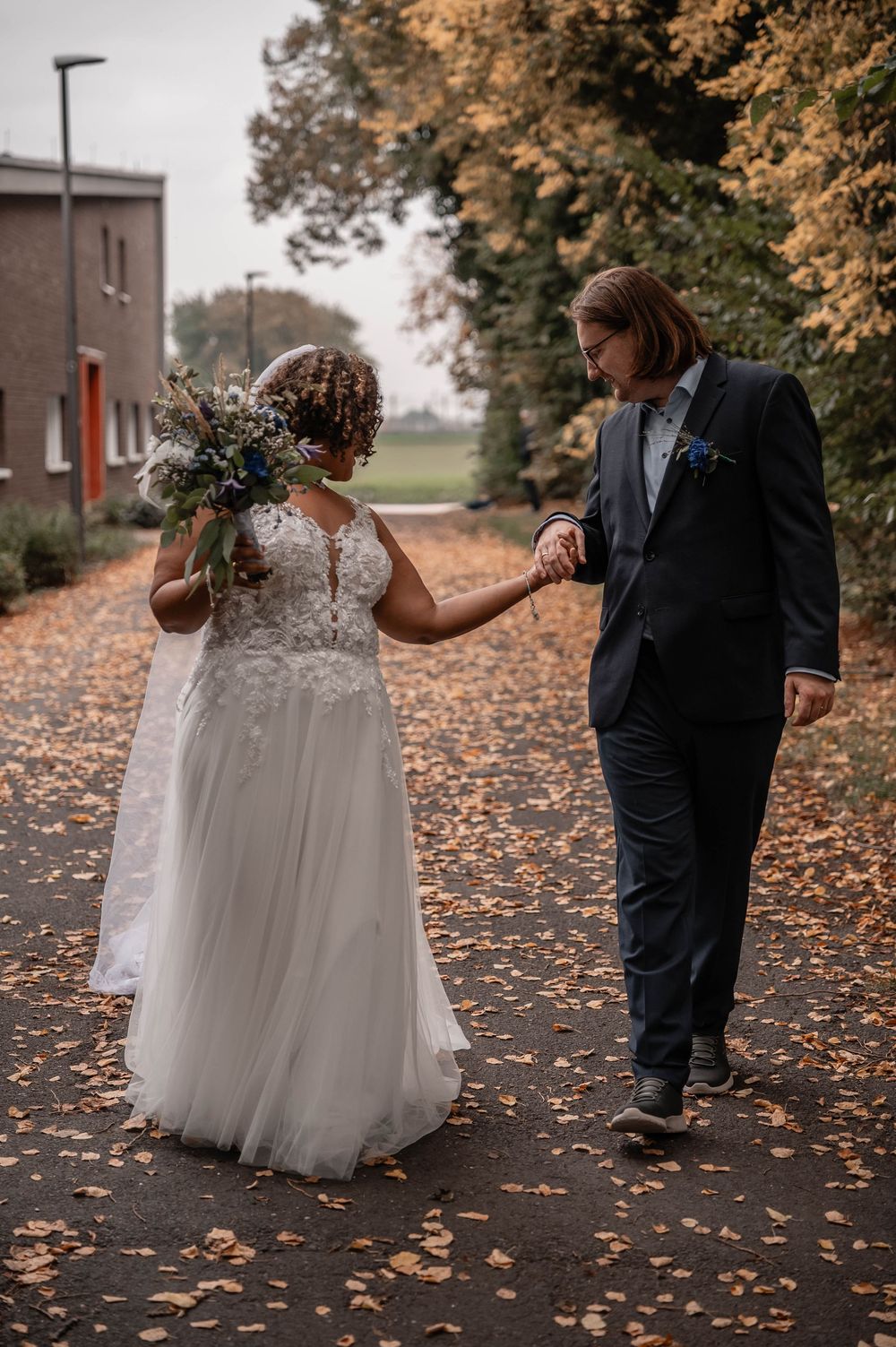 A couple in wedding attire walking on an autumn path covered with fallen leaves during their fall wedding.