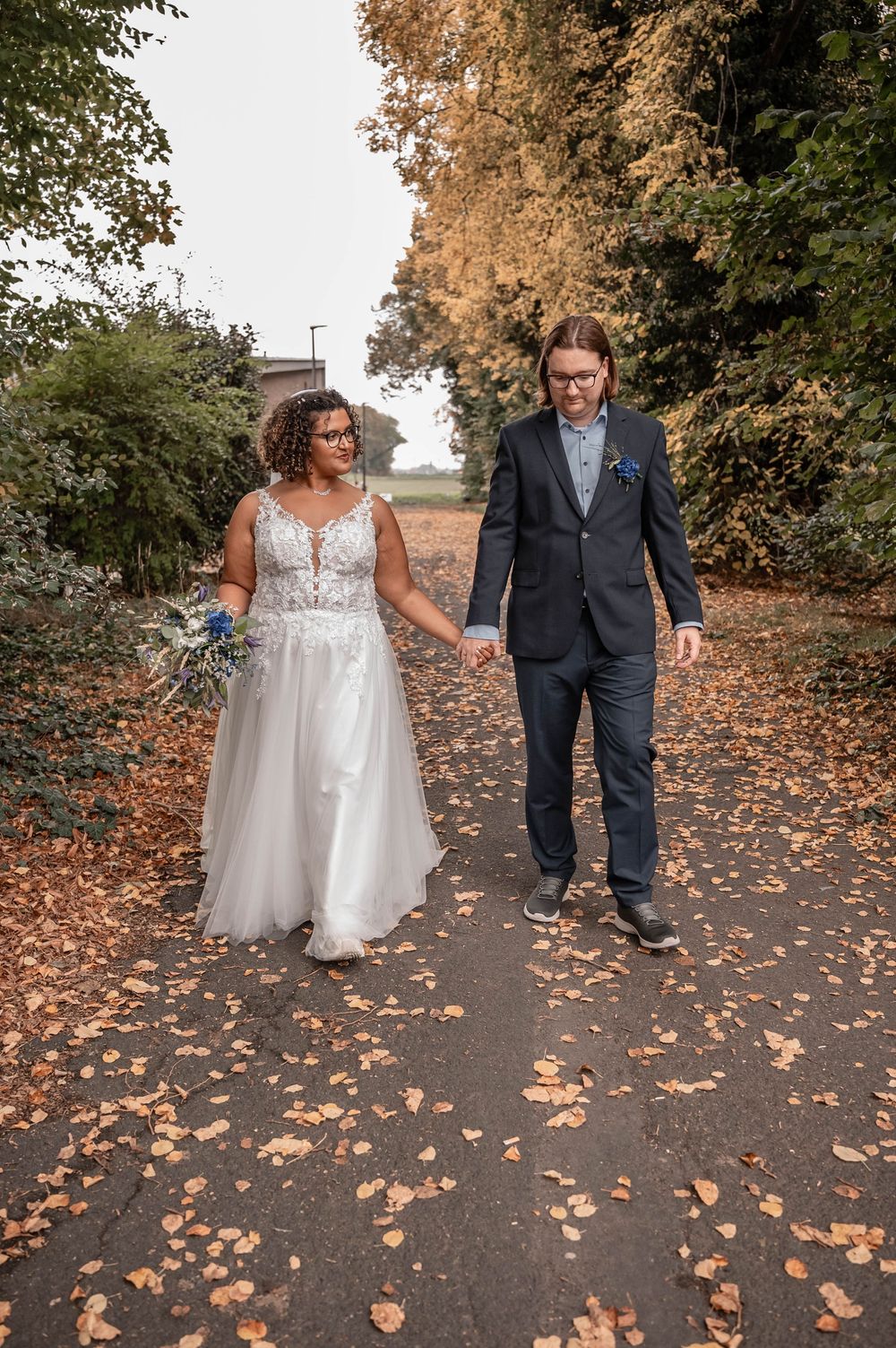 A couple in wedding attire walks along a path covered in fallen autumn leaves during an outdoor photo session.