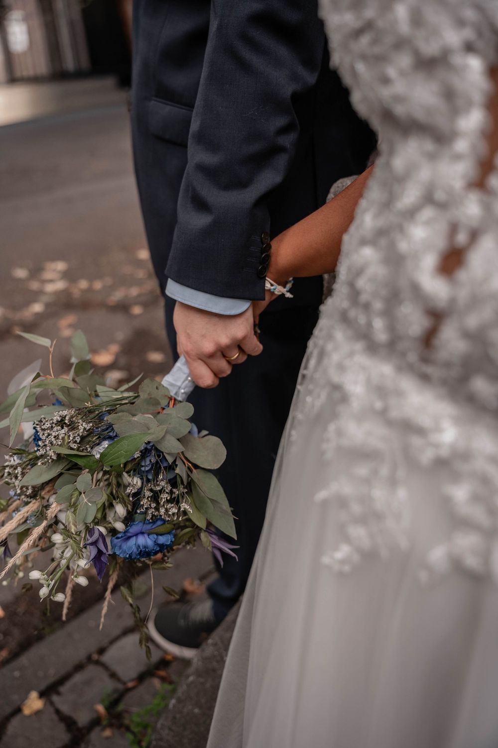 Close-up detail of wedding attire showing hands holding delicate floral bouquet with blue and purple blooms.