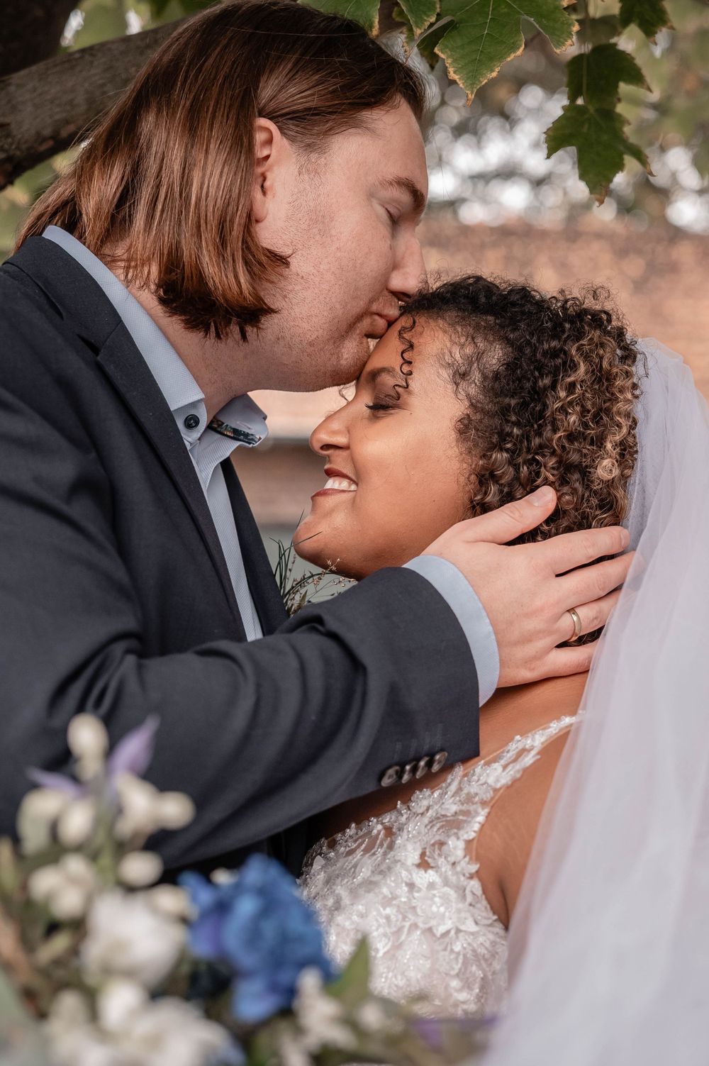 Intimate wedding portrait framed by green foliage and blue flowers showing tender embrace.