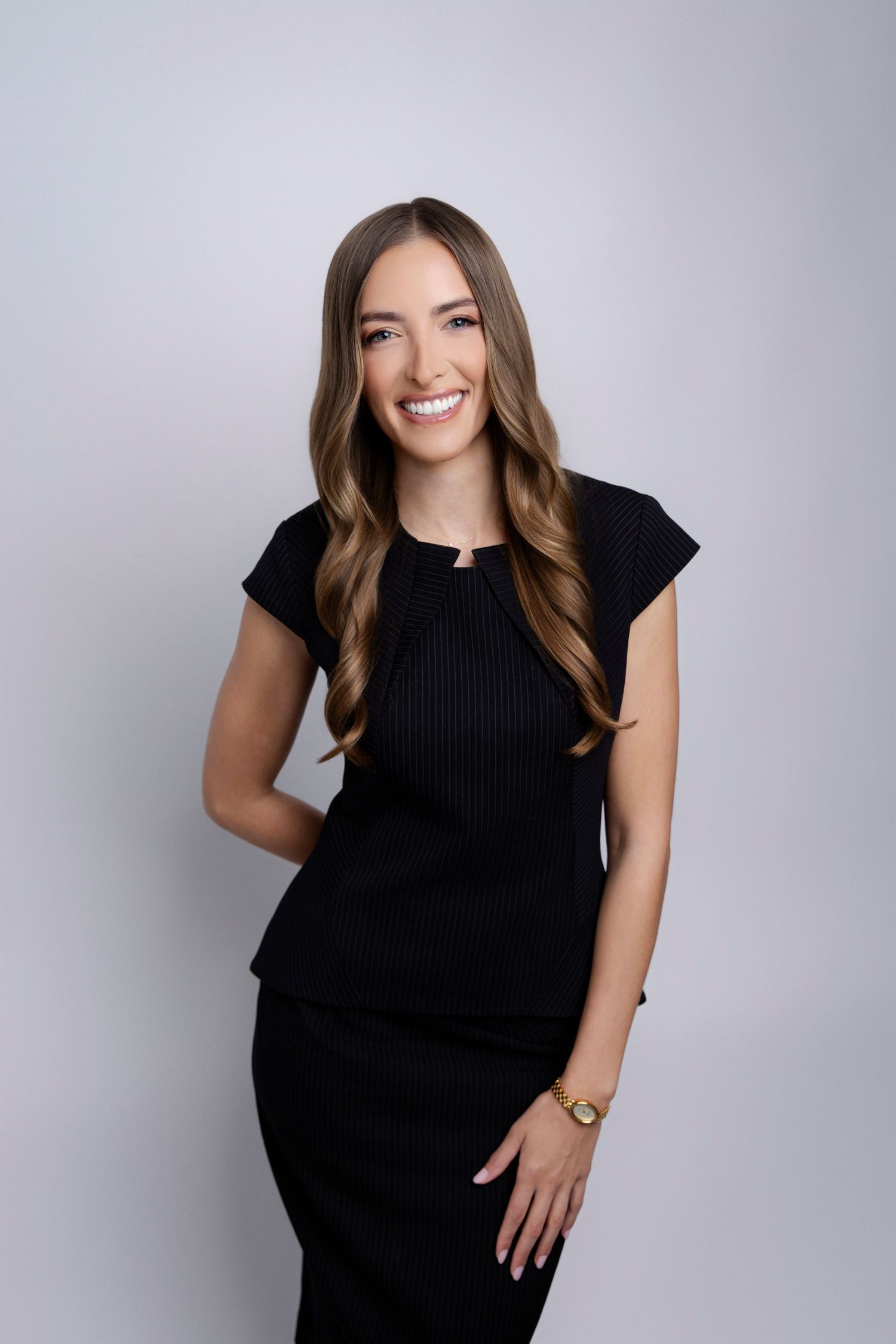 Professional studio portrait of a smiling person with long wavy brown hair wearing a black dress against a gray background.
