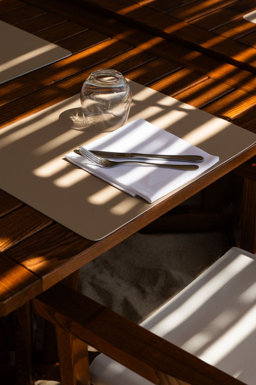Sunlit wooden restaurant table set with a placemat, white napkin, silverware, and a glass.