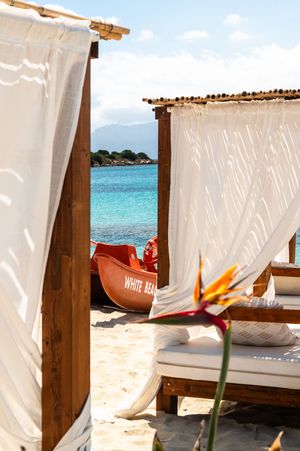 Luxury beach cabana with white curtains, sun loungers, and a red White Beach boat by turquoise waters.