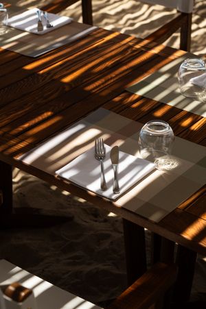Wooden restaurant table set with fork, knife, placemat, and glass in warm sunlight on sandy beach.