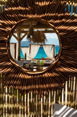 Tropical beach cabanas viewed through a circular woven rattan frame with turquoise ocean in the background.