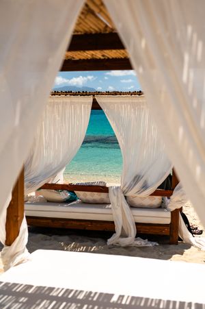 Luxurious white curtained beach cabana with wooden daybed overlooking turquoise Caribbean ocean waters.