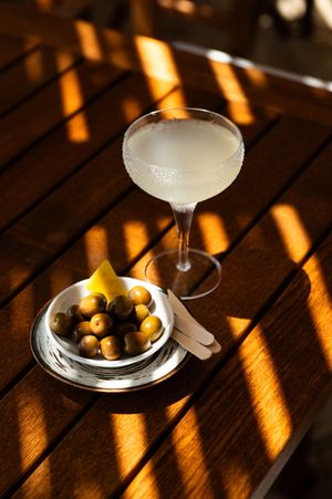 Frosted coupe cocktail glass beside a silver bowl of green olives on a sunlit wooden table.