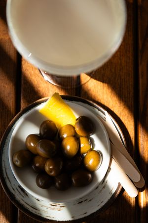A small white rustic plate with green olives and a lemon wedge beside a white drink on a wooden table.