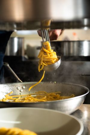 Chef twirling fresh yellow egg pasta with a fork over a steaming pan in a professional kitchen.