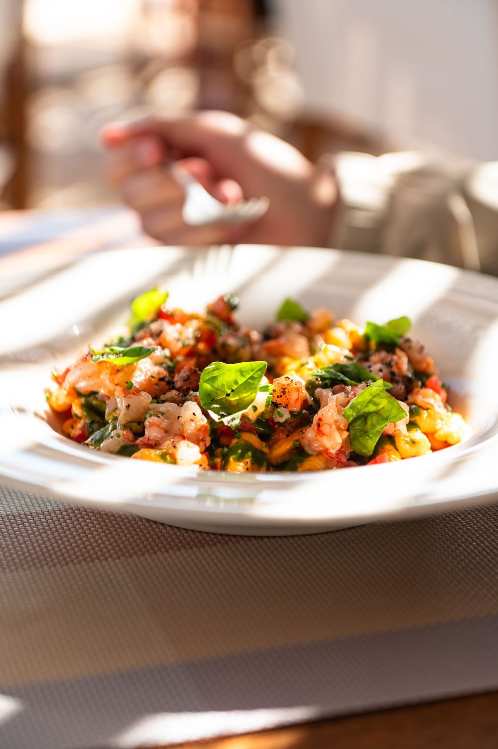 A white plate of pasta with tomato sauce, ground meat, and fresh basil leaves served in a sunlit restaurant.