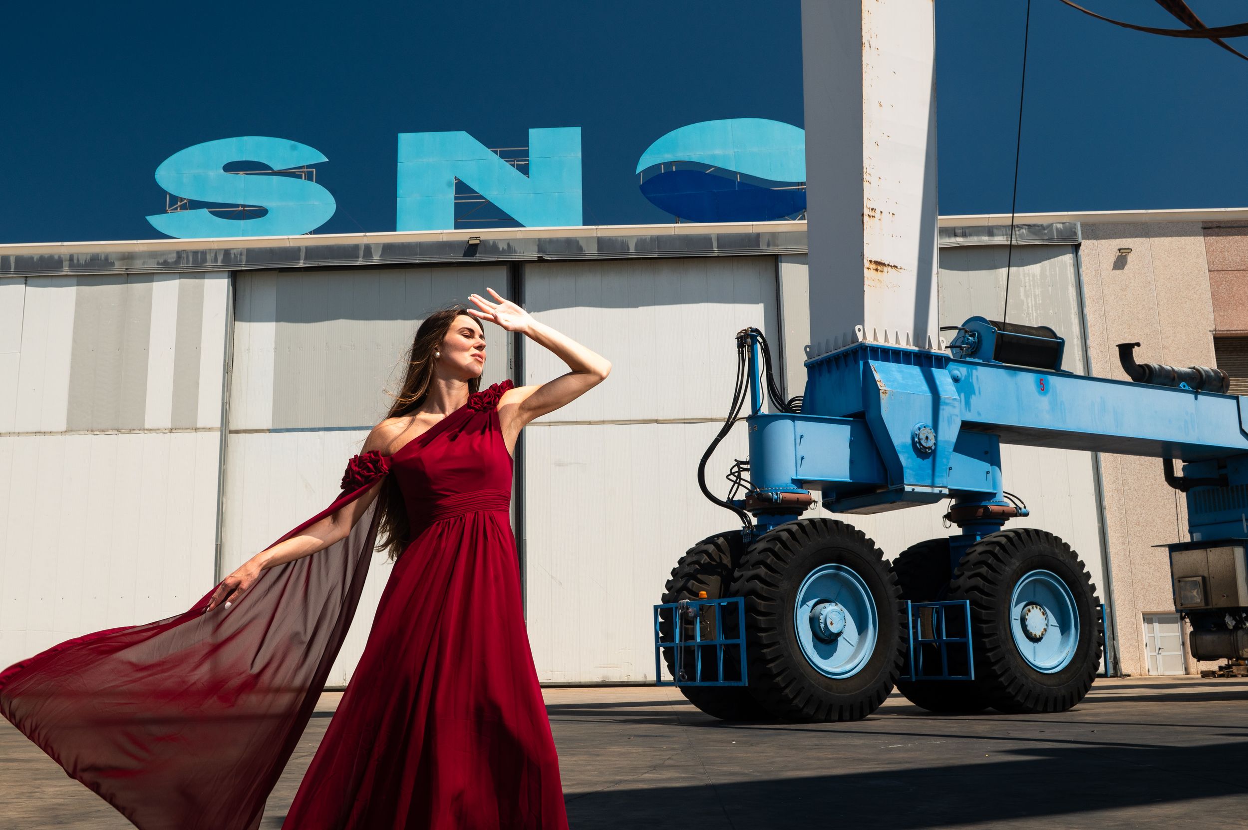 Woman in red gown poses before blue industrial crane and SNO building in dramatic outdoor fashion shoot.