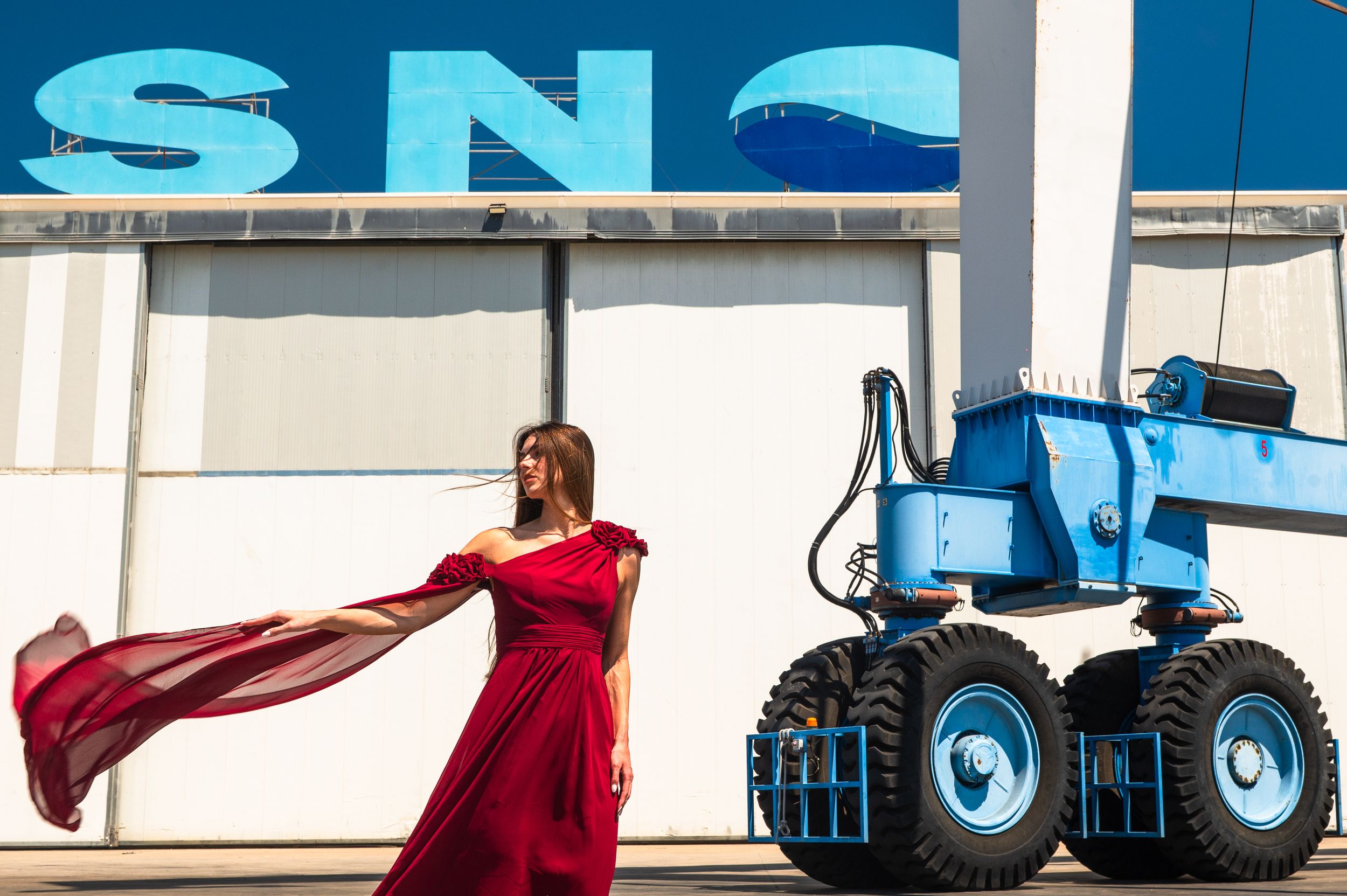 Woman in red flowing dress poses beside a blue industrial vehicle in front of a blue and white building.