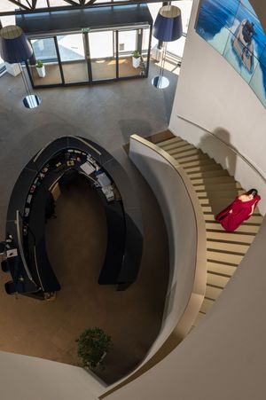Aerial view of a modern office atrium with curved staircase, circular workstation, and person in red dress descending stairs.