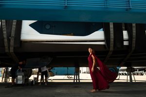 Woman in flowing red gown poses beneath a large blue structure outdoors, with workers visible in the background.