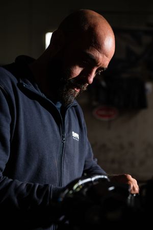 Bald bearded man in navy blue hoodie carefully inspects dark motorcycle helmet in dimly lit garage.
