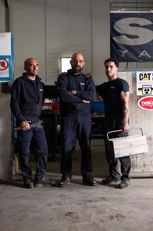 Three male mechanics in dark uniforms stand confidently in an industrial garage workshop.
