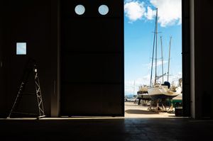 Sailboats on dry dock visible through large open hangar doors with circular windows and blue sky beyond.