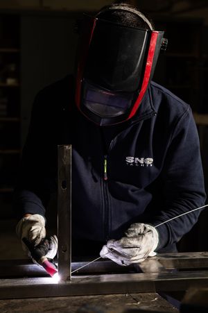 A professional welder wearing a protective mask and SNS Yachts jacket welding metal components in a workshop.