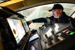 Man in cap operating controls in yellow machinery cab with digital screens.