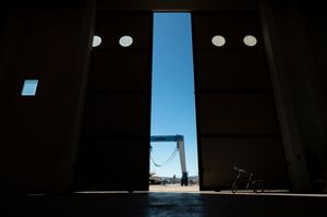 Large industrial hangar doors open to reveal a bright blue sky, with a bicycle silhouetted in the doorway.